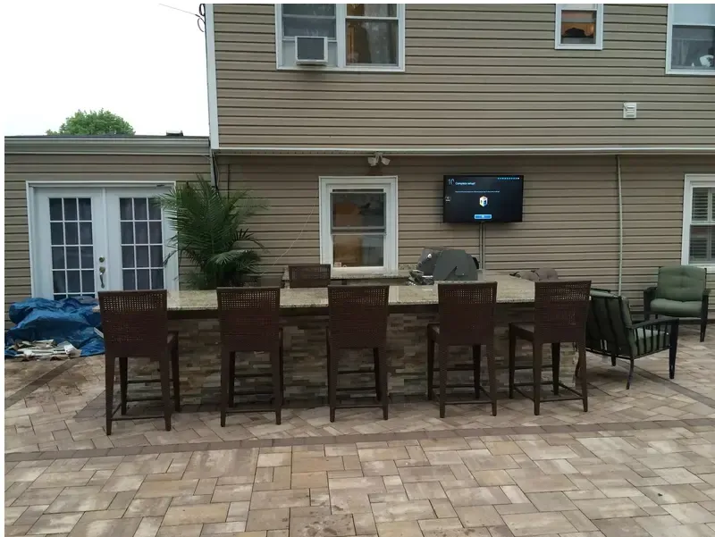 Outdoor bar with seating for eight, mounted TV on a siding-clad house, brick patio.