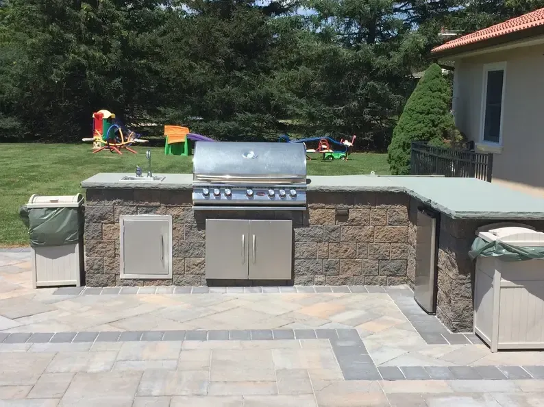 Outdoor kitchen with grill, sink, and fridge, built into a stone structure on a patio with a playground in the background.