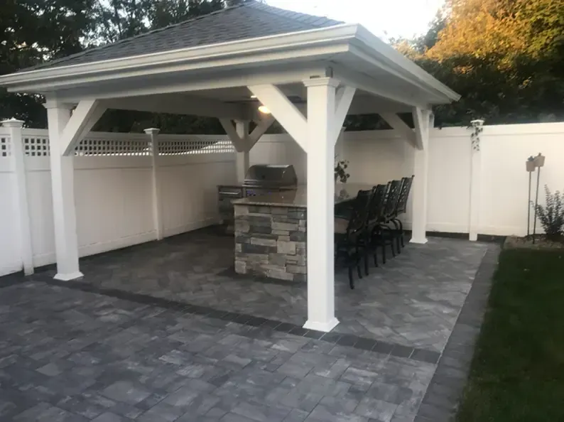Outdoor pavilion with gray pavers, built-in grill, white walls, fence, and a row of black chairs.