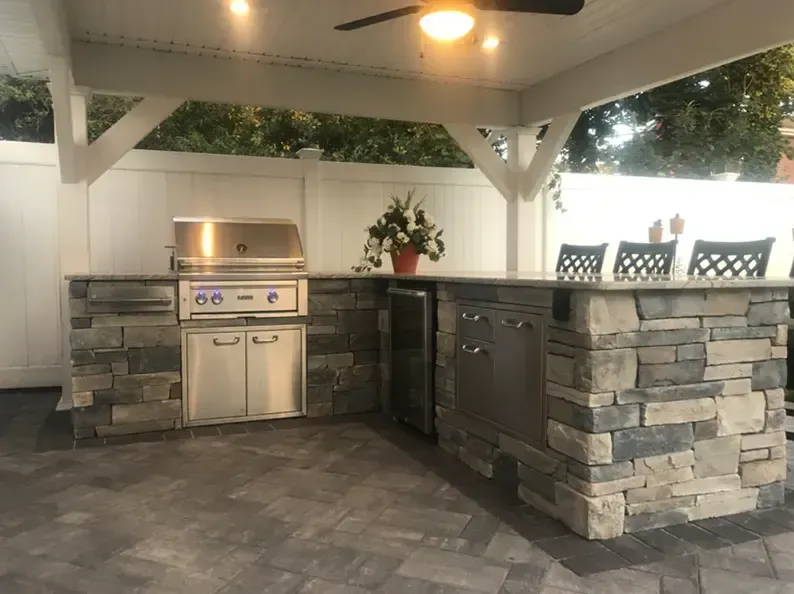 Outdoor kitchen with stone-clad counters, grill, refrigerator, and seating area under a white pergola, and fence.