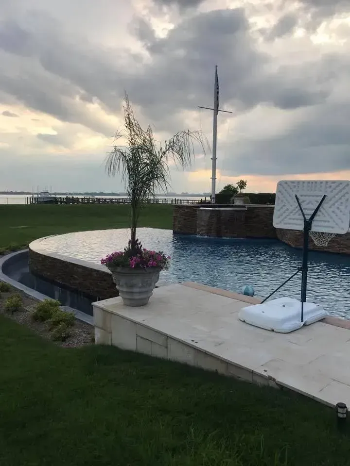 Poolside view: Pool with waterfall, basketball hoop, potted flowers, grassy lawn, water view, cloudy sky.
