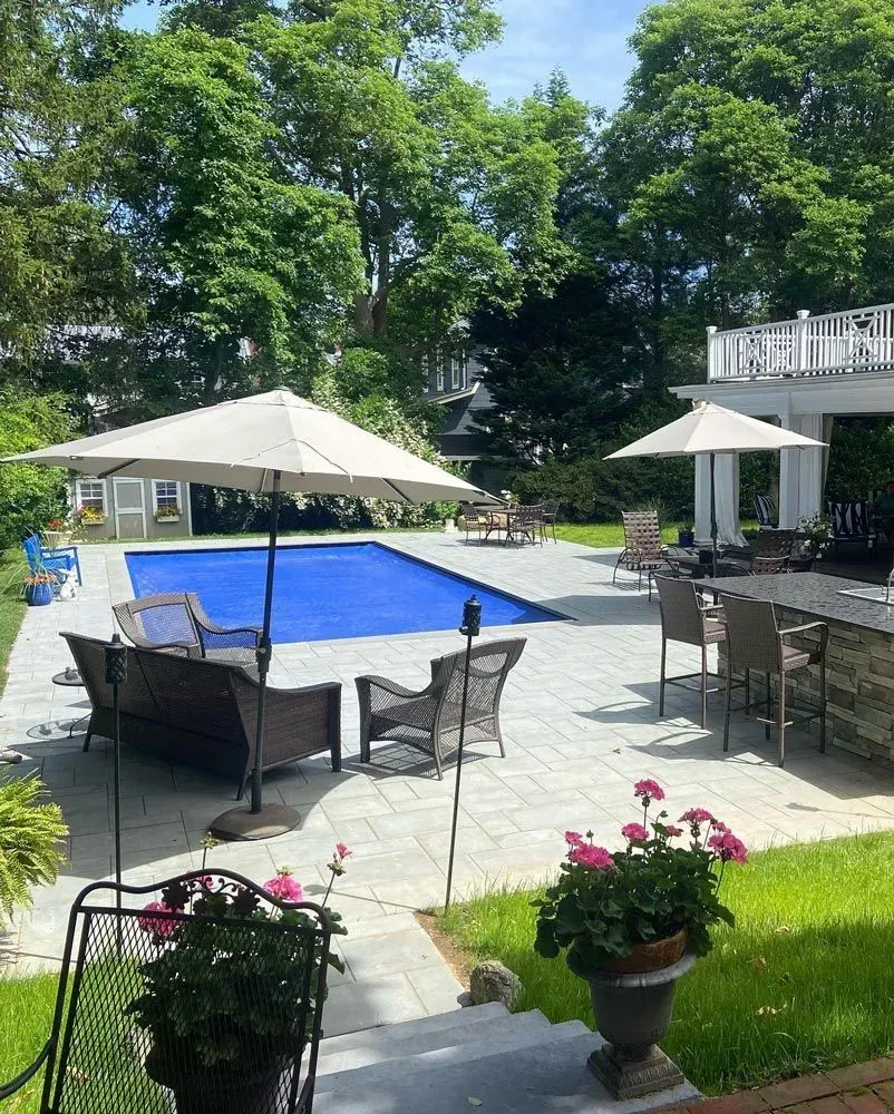Patio with pool, umbrellas, seating, and greenery. Blue pool cover, light tan umbrellas, and stone patio.