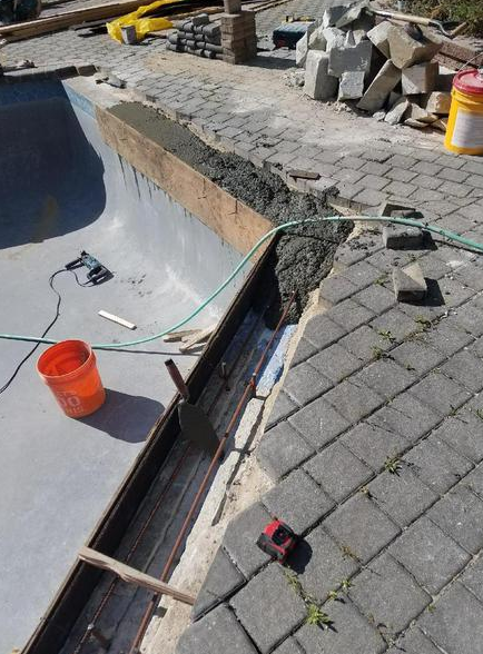 Construction of a pool coping. Concrete being poured between the pool and brick patio. Tools and materials visible.