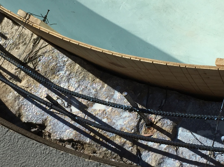 Reinforcement bars inside curved wooden mold for concrete construction. Sunlight, gray tones.