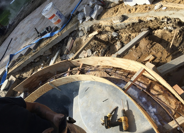 Construction of a curved concrete structure with wooden forms, rebar, and tools in a dirt setting.
