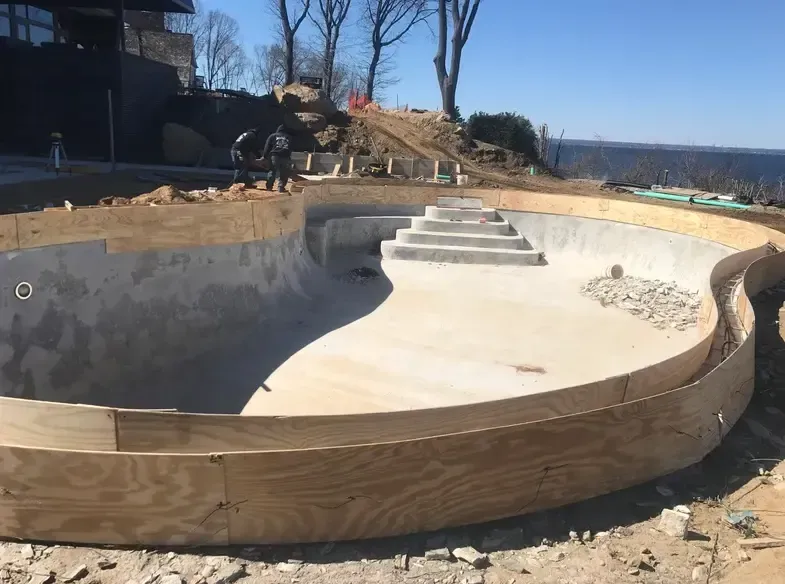 Construction of an empty, curved swimming pool with steps. Wooden formwork surrounds the concrete structure.
