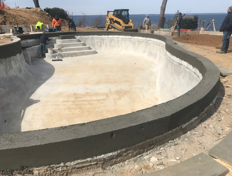 A concrete pool under construction with steps, surrounded by a concrete border, at a construction site overlooking the ocean.