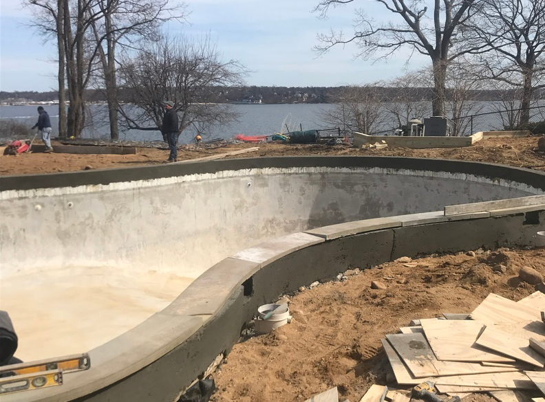 Construction of a concrete pool near a body of water, two workers visible, sunny day.