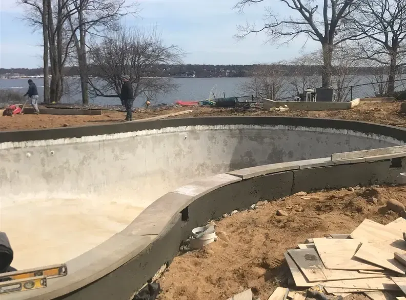 Pool construction in progress, concrete shell, workers, waterfront setting.