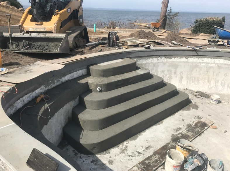 Concrete pool construction: Steps leading into the pool, heavy machinery, coastal backdrop.