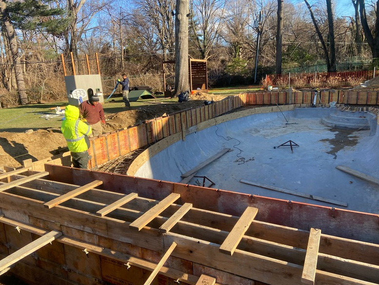 Pool construction site. Workers near wooden forms. Trees and grass in the background.