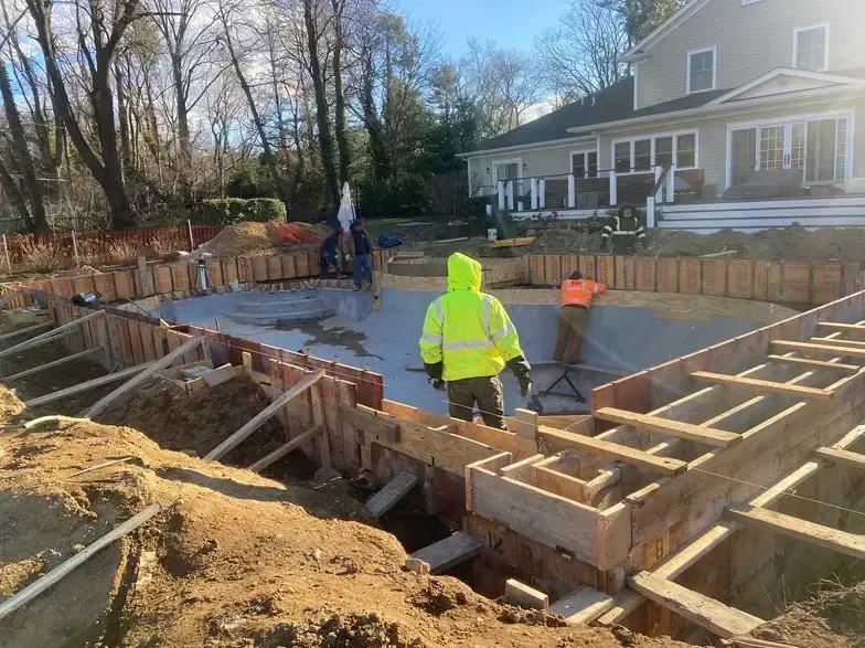 Construction site with workers pouring concrete into a pool form. Yellow safety vests, brown wood forms.