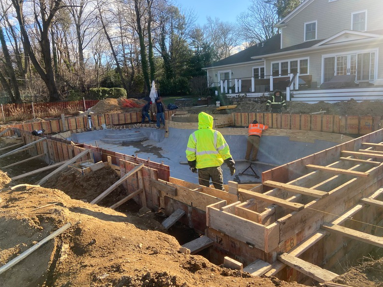 Construction of a swimming pool. Workers stand near wooden forms. Dirt surrounds the excavation.