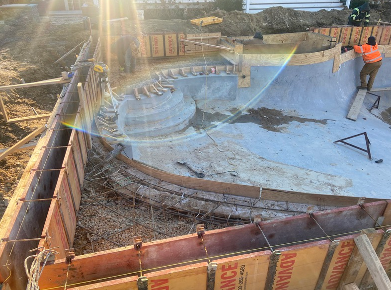 Construction site with a pool-like form, workers, wooden forms, and concrete elements.