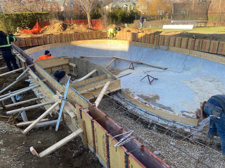 Construction site of a pool. Workers build forms for concrete; earthmoving equipment visible in background.