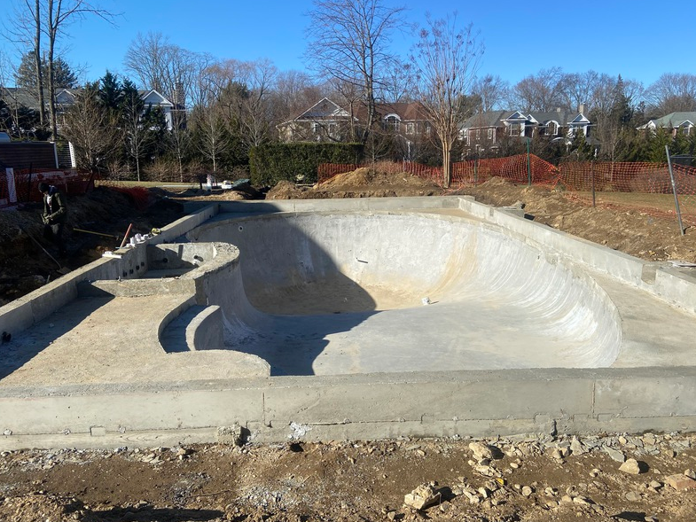 Unfinished concrete skatepark bowl under construction, with exposed dirt and surrounding fencing; clear, sunny day.
