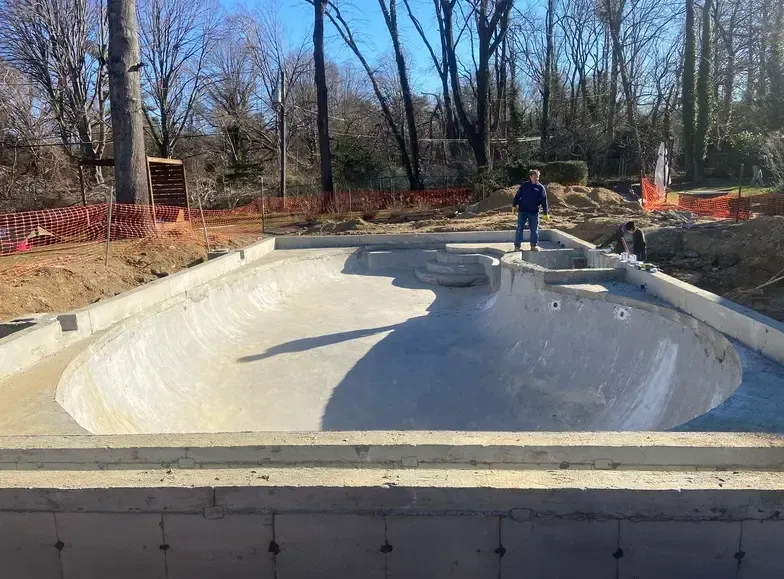 Concrete skate bowl under construction; person standing at the edge of the bowl, surrounded by dirt and fencing.