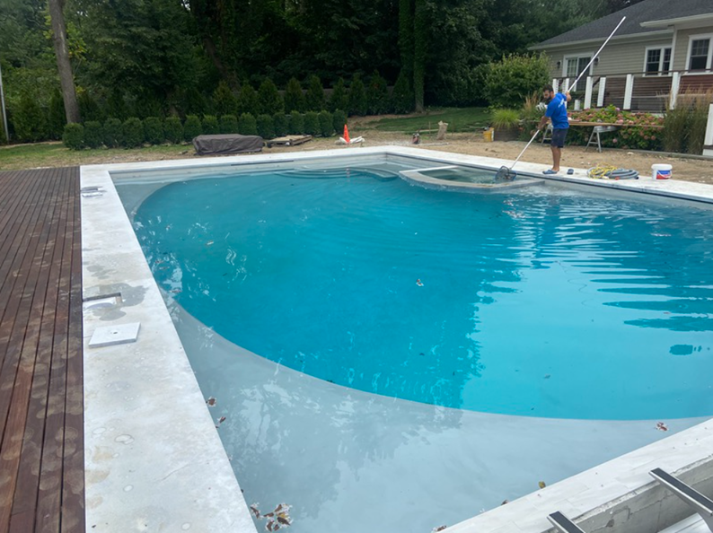 A pool with a person cleaning it, surrounded by a deck and trees. Blue water, gray concrete.