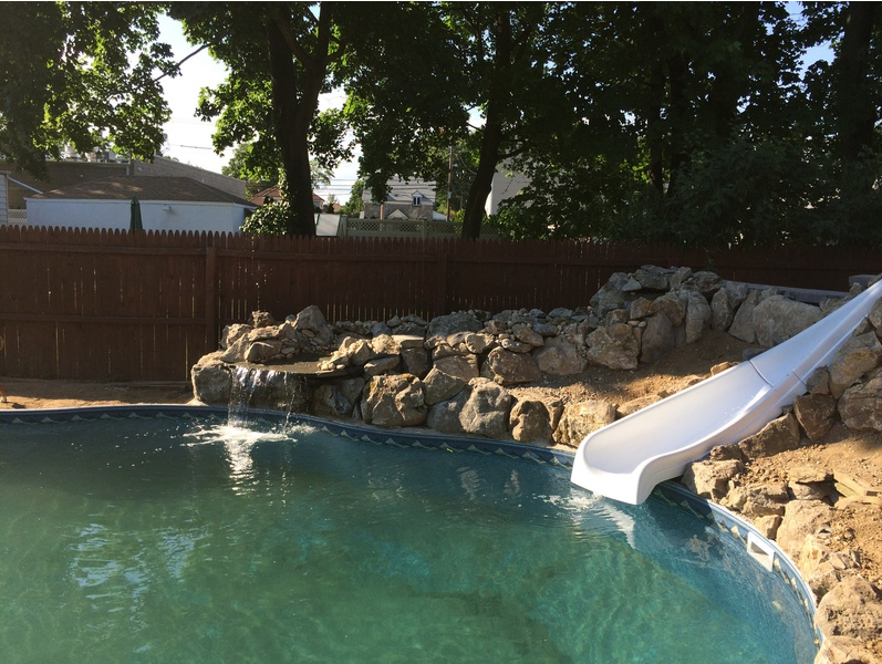 A pool with a waterslide, waterfall, and rock landscaping in a backyard setting.