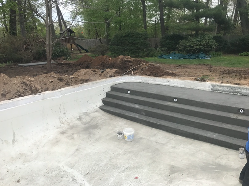 Concrete pool under construction with steps and lights; dirt and trees in background.
