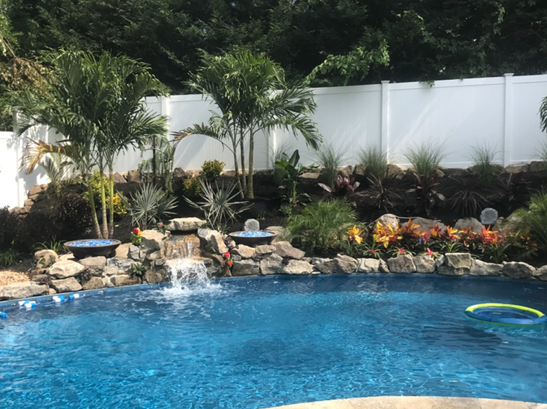 Backyard pool with waterfall, surrounded by lush landscaping and a white privacy fence.