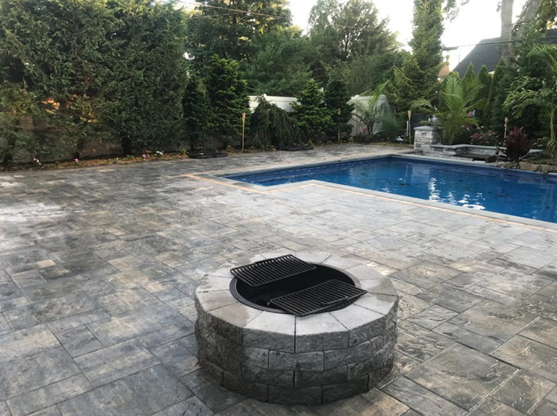 Fire pit and pool in a backyard with a paved patio and surrounding greenery.
