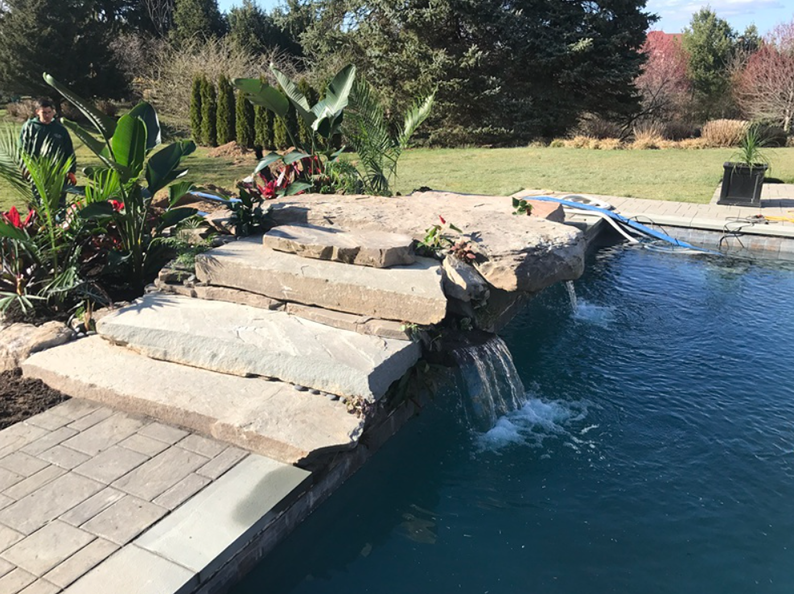 Stone waterfall cascades into a pool, steps lead up to a rock ledge with tropical plants, green lawn in background.