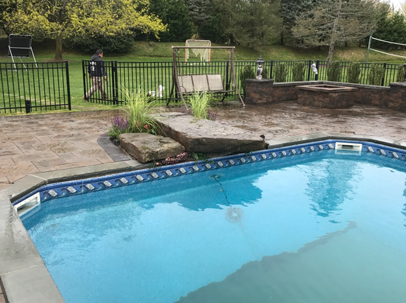 Poolside scene: a blue-tiled pool, patio, black fence, swing, person walking, and foliage.
