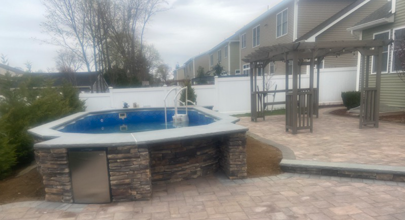A backyard with a small blue pool, stone surround, and a wooden pergola.