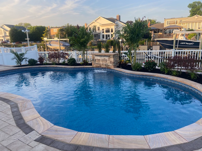 Pool with water feature, stone patio, and waterfront houses in the background.