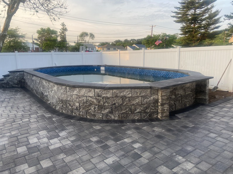 Oval-shaped swimming pool with stone walls and a gray paver patio. A white fence and trees are in the background.
