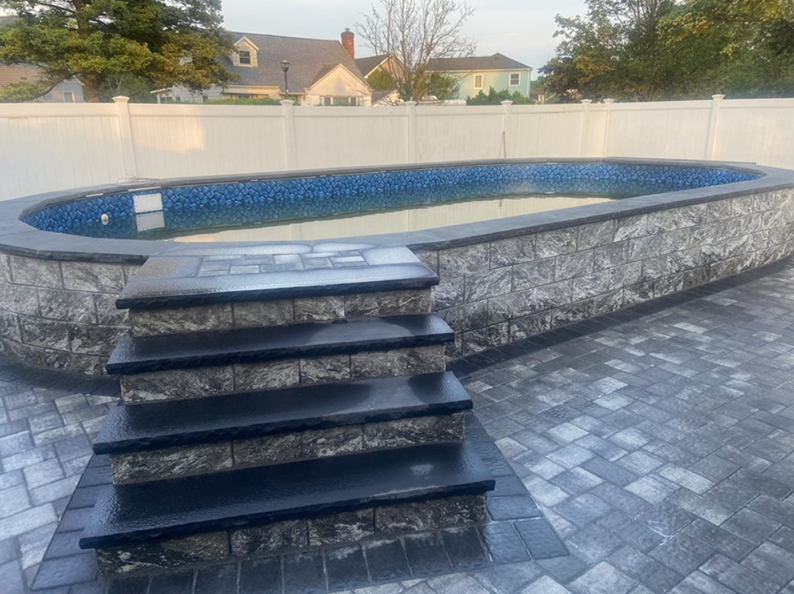 Above-ground pool with steps and a gray stone facade. Houses and a white fence are in the background.