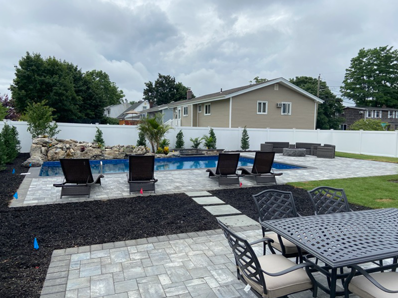 Backyard with a pool, lounge chairs, and dining area. Gray pavers and black mulch. House and white fence in the background.