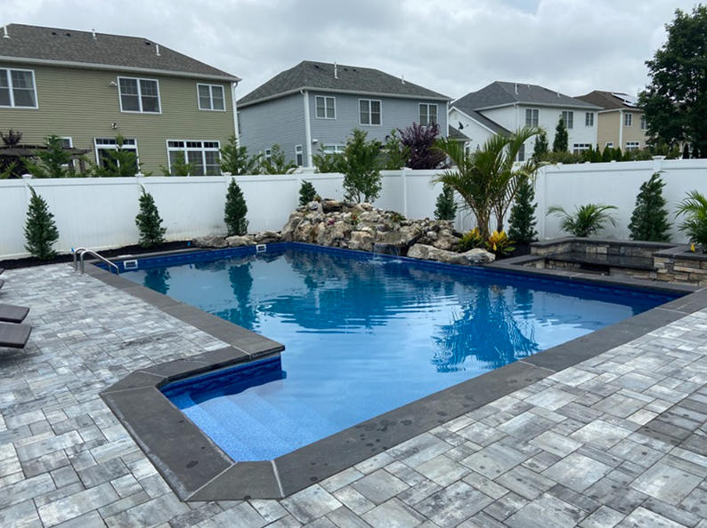 Backyard pool with waterfall feature and patio. Houses and cloudy sky in the background.