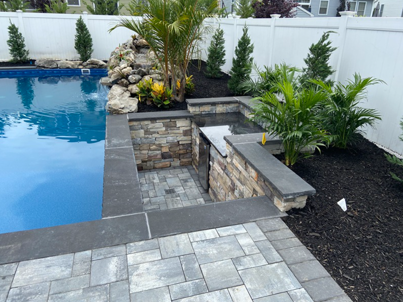 A pool with a waterfall, stone seating, and palm trees against a white fence.