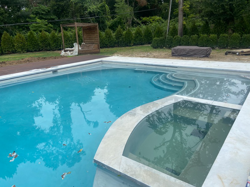 Swimming pool with attached spa and steps. Blue water, gray concrete, and surrounding greenery.