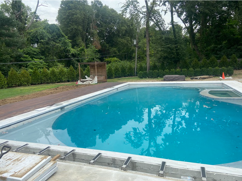 A partially finished swimming pool surrounded by greenery and a wooden deck on a cloudy day.