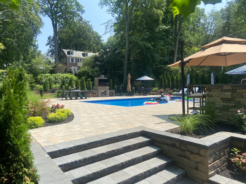 Backyard with pool, patio, steps, and trees; umbrellas and seating. Sunny day.