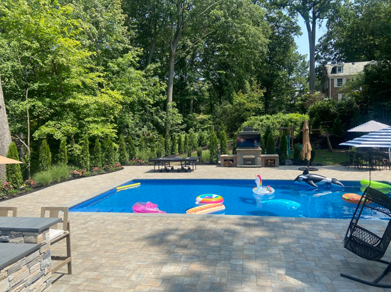 Backyard pool with inflatables. Blue water, stone patio, grill area, trees, and a swing chair.