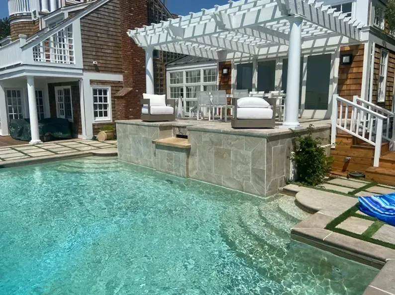 Pool with steps, a pergola, and seating area, in front of a house on a sunny day.