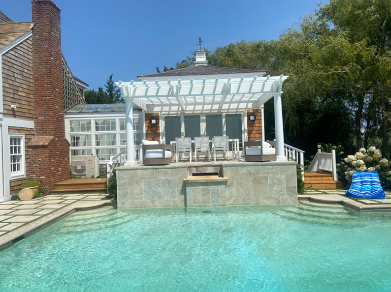 Poolside pergola with seating, near a brick building. Light blue water and clear sky.