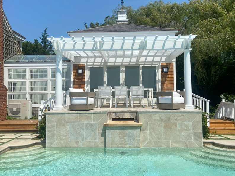 Outdoor dining area with white pergola over a pool. Brown stone wall.