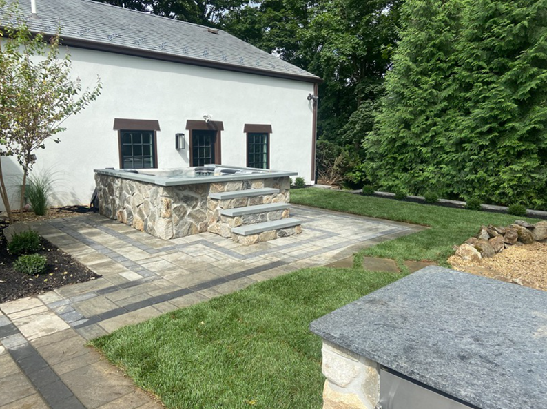 Stone hot tub with steps, set in a paved patio area beside a white building and green lawn.