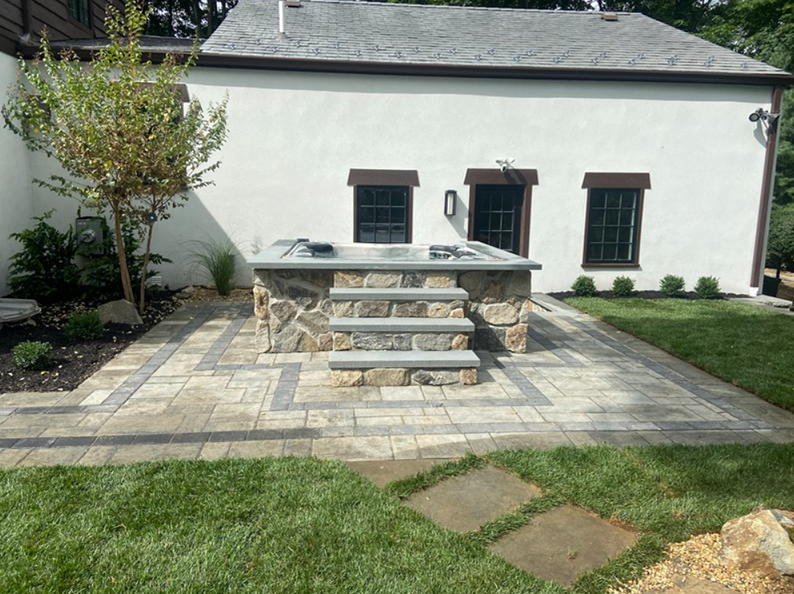 Stone hot tub with steps on a paved patio, against a white building, grass and landscaping.