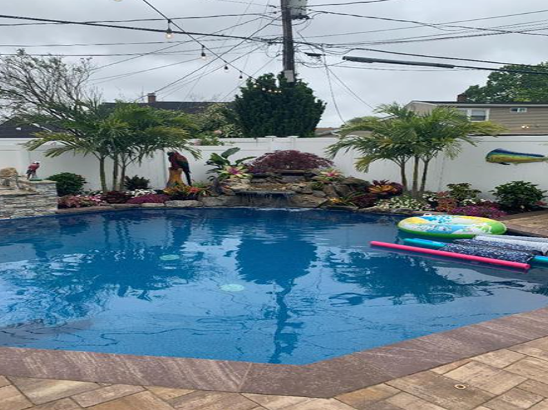 Pool with waterfall, palm trees, and colorful inflatables in a backyard on an overcast day.