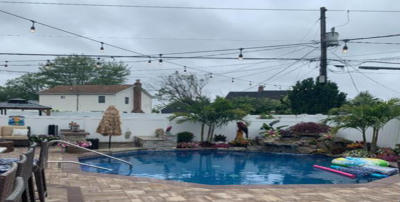 Swimming pool in backyard on a cloudy day, with string lights, white walls, and lush landscaping.