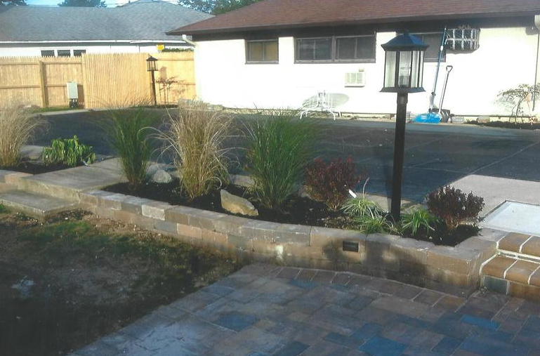 A brick planter bed with ornamental grasses, a lamppost, and a paved driveway and patio in the backyard.