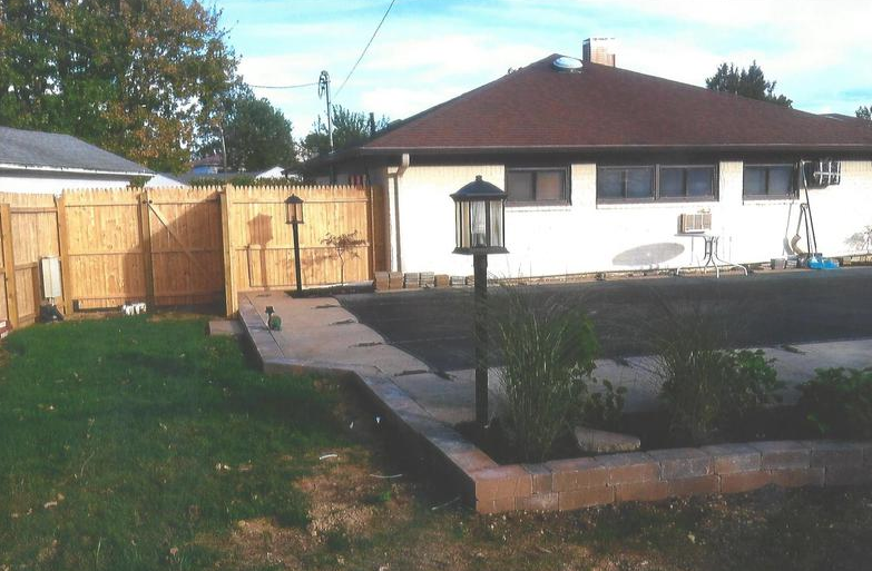 A house with a brown roof and a wooden fence in front. Two street lights and a paved path.