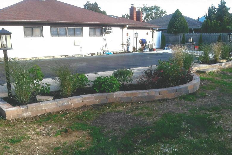 Backyard with a brick-edged garden bed, paved area, and a house. Someone is working in the garden.