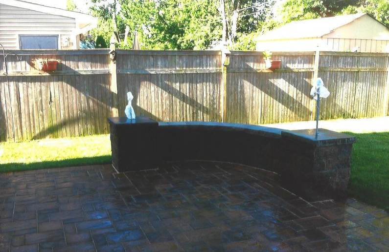 A curved stone bench on a brick patio in front of a wooden fence.